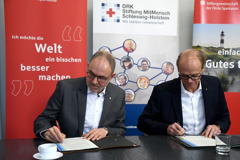 Ralph Schmieder (DRK) und Carsten Zarp (Förde Sparkasse) sitzen nebeneinander an einem Tisch und unterzeichnen Verträge.